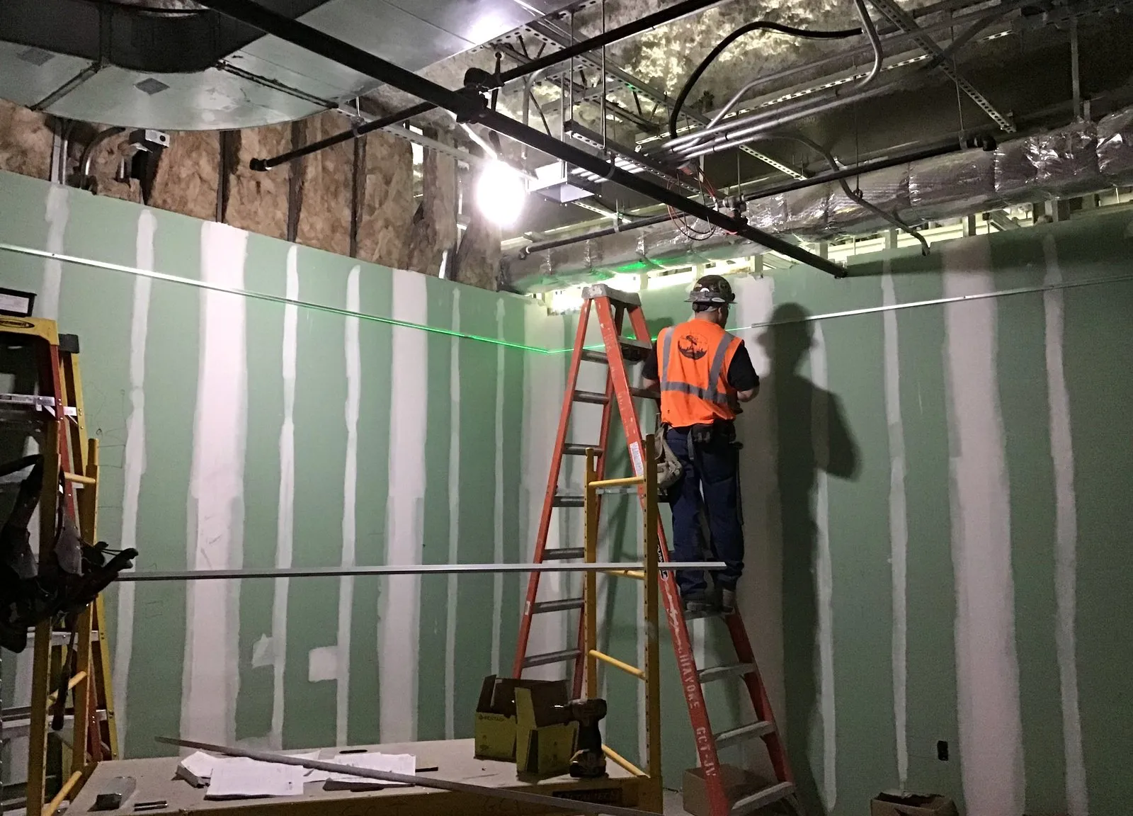A fitter on a stepladder fixing the wall trim for a suspended ceiling, with a green laser line across the boarded wall
